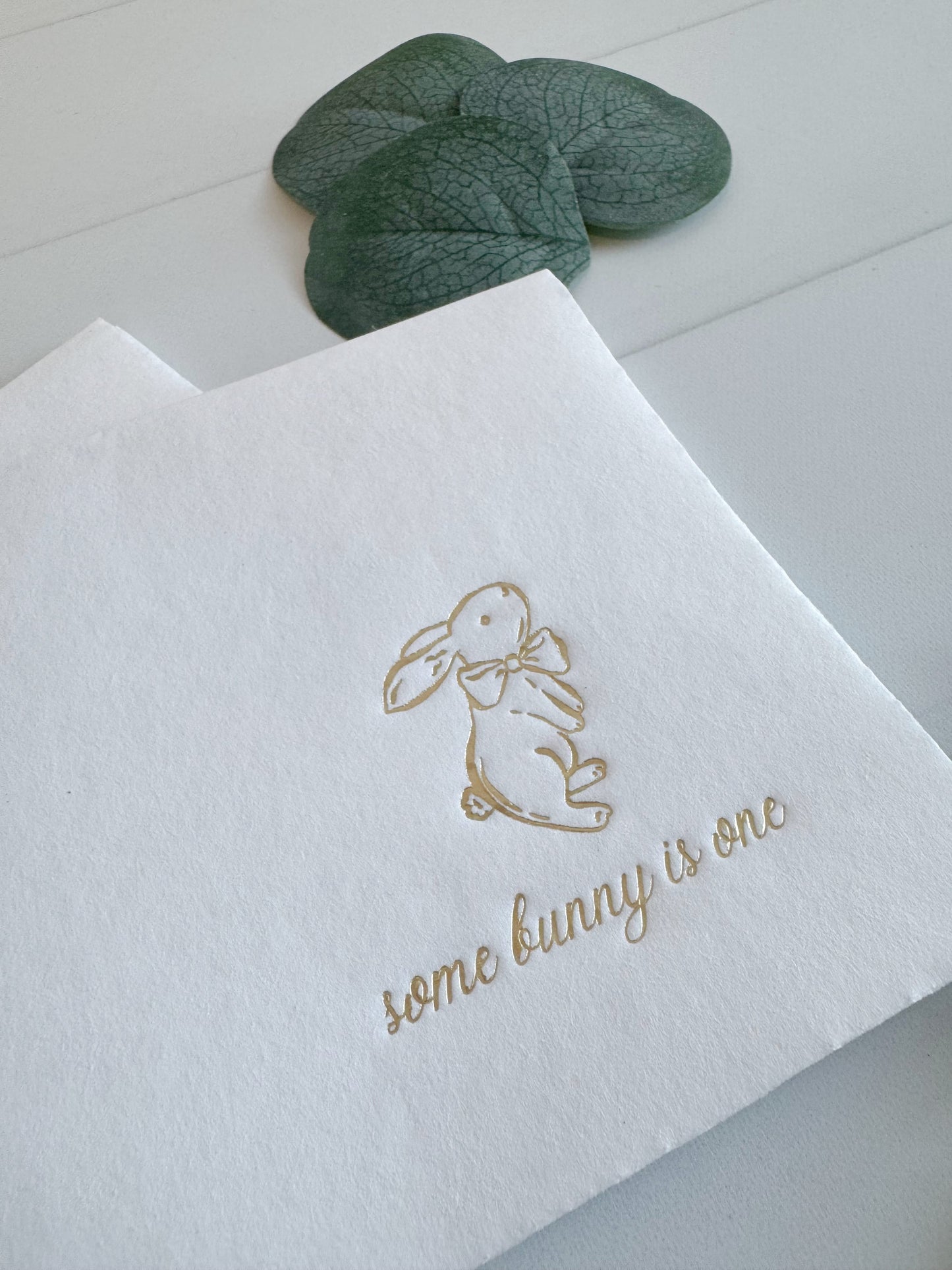 White paper napkins with a gold foil bunny illustration and the phrase 'some bunny is one' in cursive, styled on a white surface with green leaves in the background.
