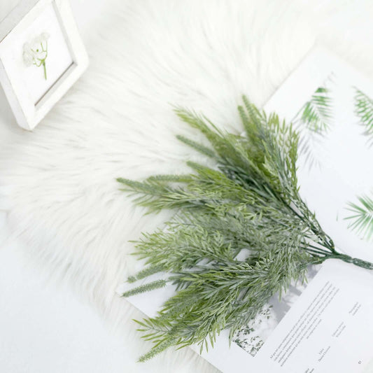 Artificial green sagebrush fern branches with a frosted, dusty-silver sheen, arranged on a white faux fur surface next to an open book and a small white frame.