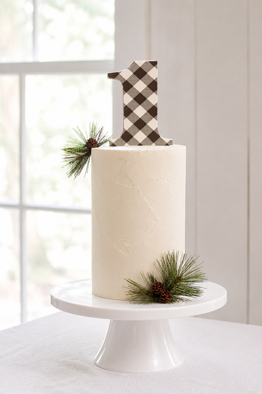 A white frosted birthday cake on a white pedestal stand, decorated with a bback and white buffalo plaid number 1 topper and pine sprigs for a lumberjack first birthday