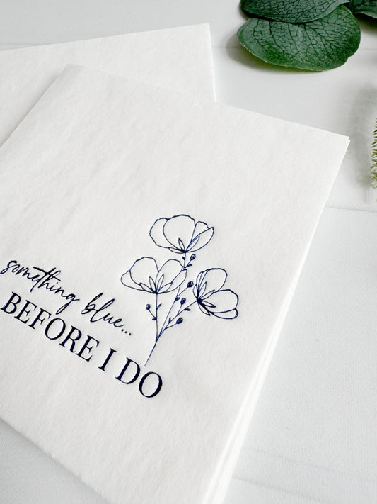 White designer cocktail napkin with floral design and text 'something blue... BEFORE I DO' on a white background.