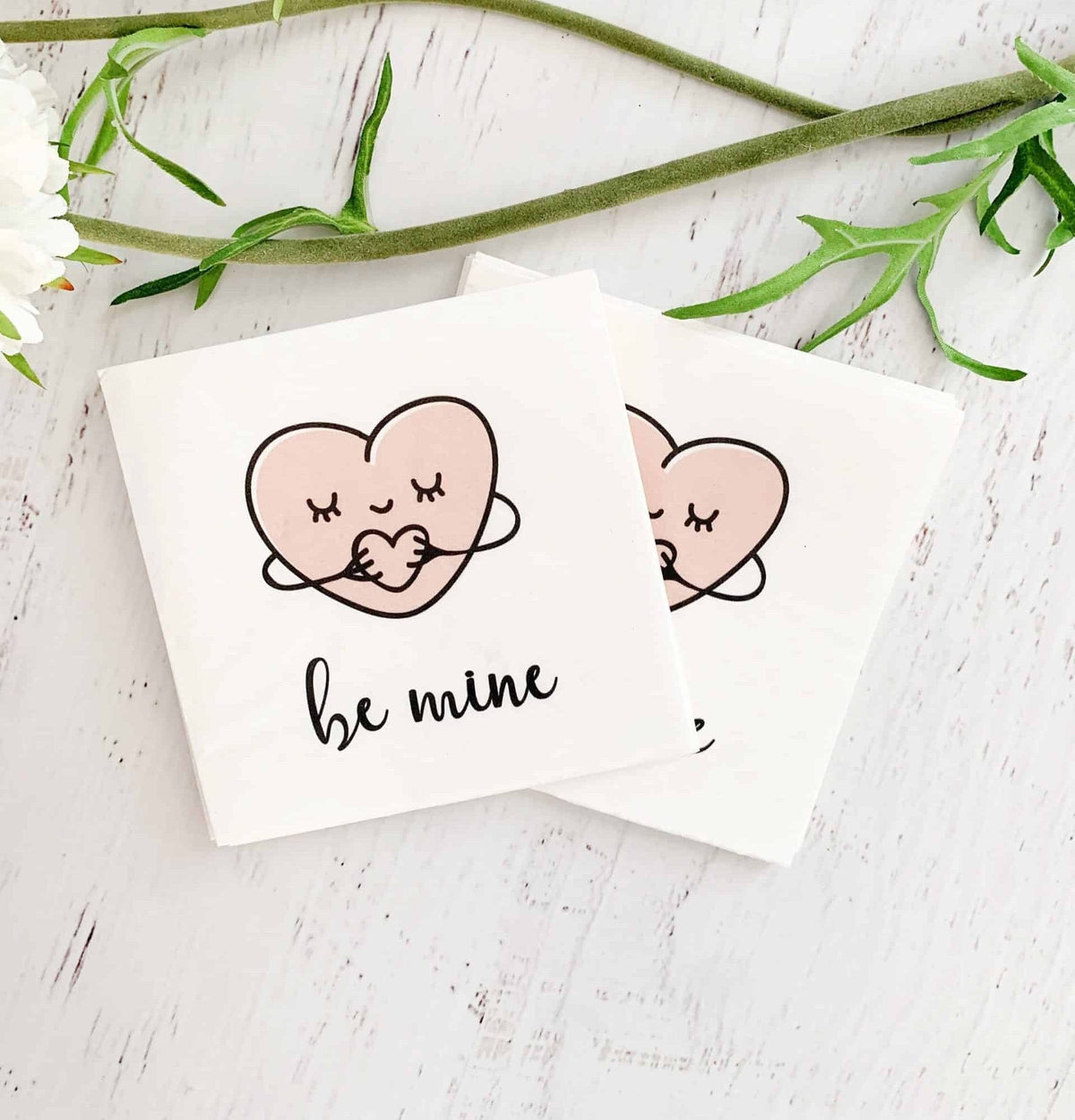 Sweet hugging hearts say "be mine" on these EnFete original cocktail napkins. White background with blush heart and black writing. Perfect for a Valentine treat, a Galentine's day cocktail, or under an afternoon snack.