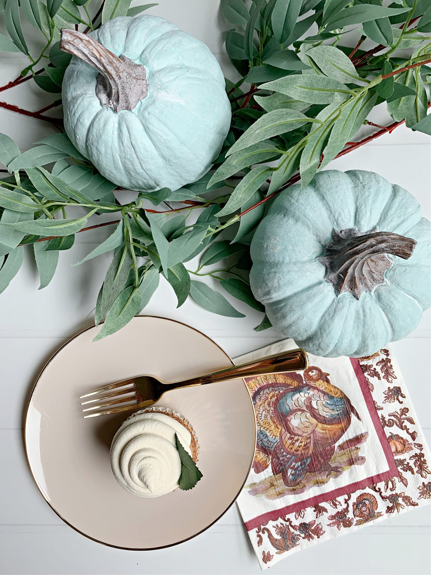Two light blue pumpkins with green leaves on a white surface, next to a plate with a dessert and a decorative napkin.