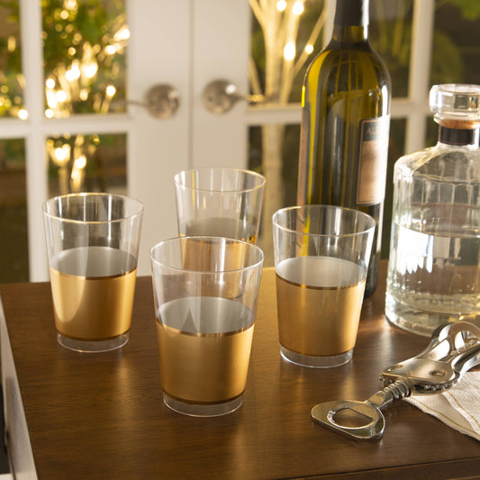 Clear plastic glasses with gold bands on a wooden table with a bottle and carafe in the background.