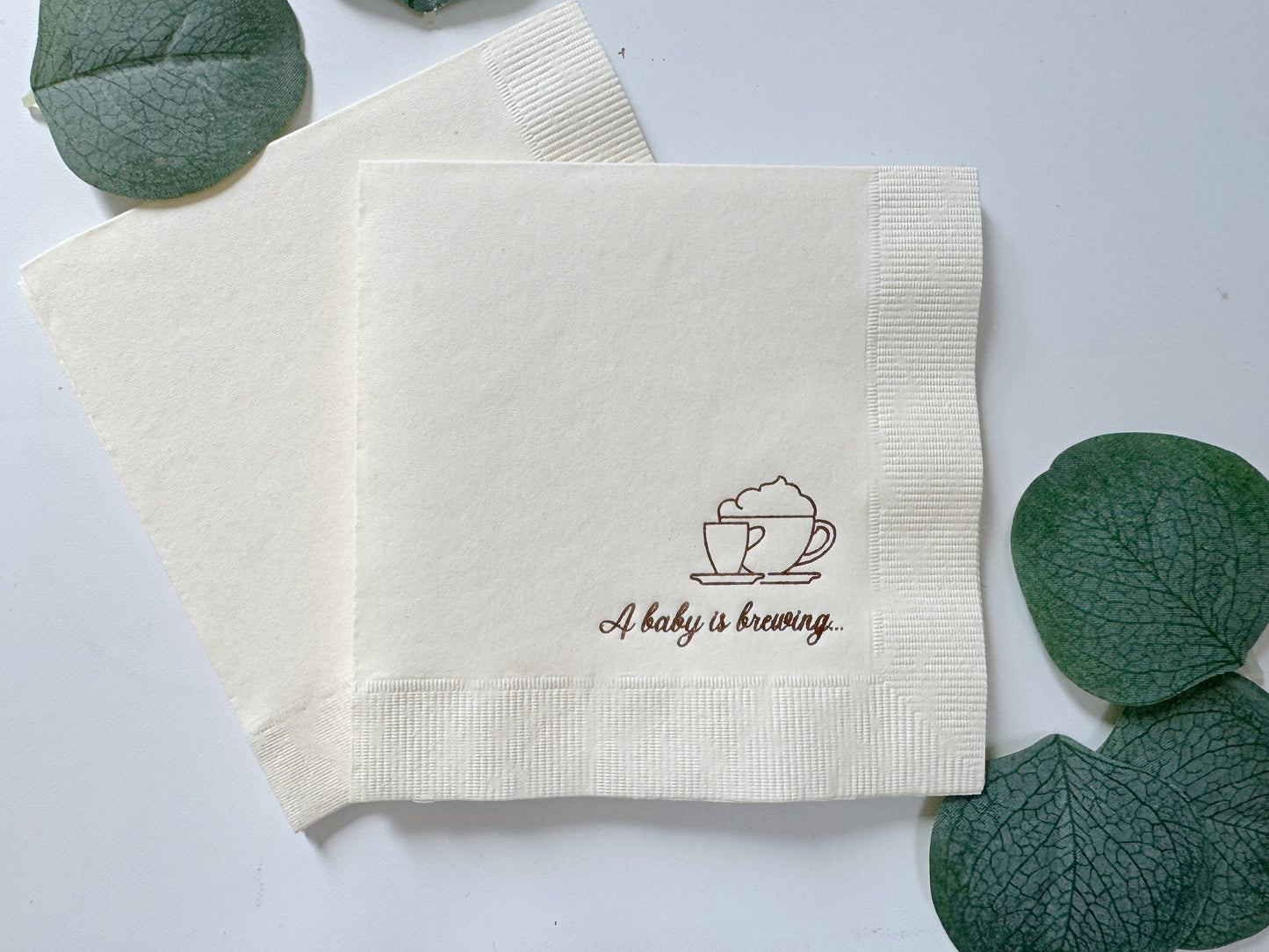 Napkin with 'A baby is brewing' text and coffee cup design on a white background with green leaves.