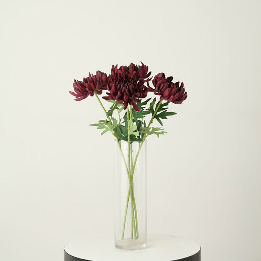 A bouquet of deep burgundy chrysanthemums with green stems and leaves arranged in a clear cylindrical glass vase, set on a round white and black pedestal against a plain off-white background.