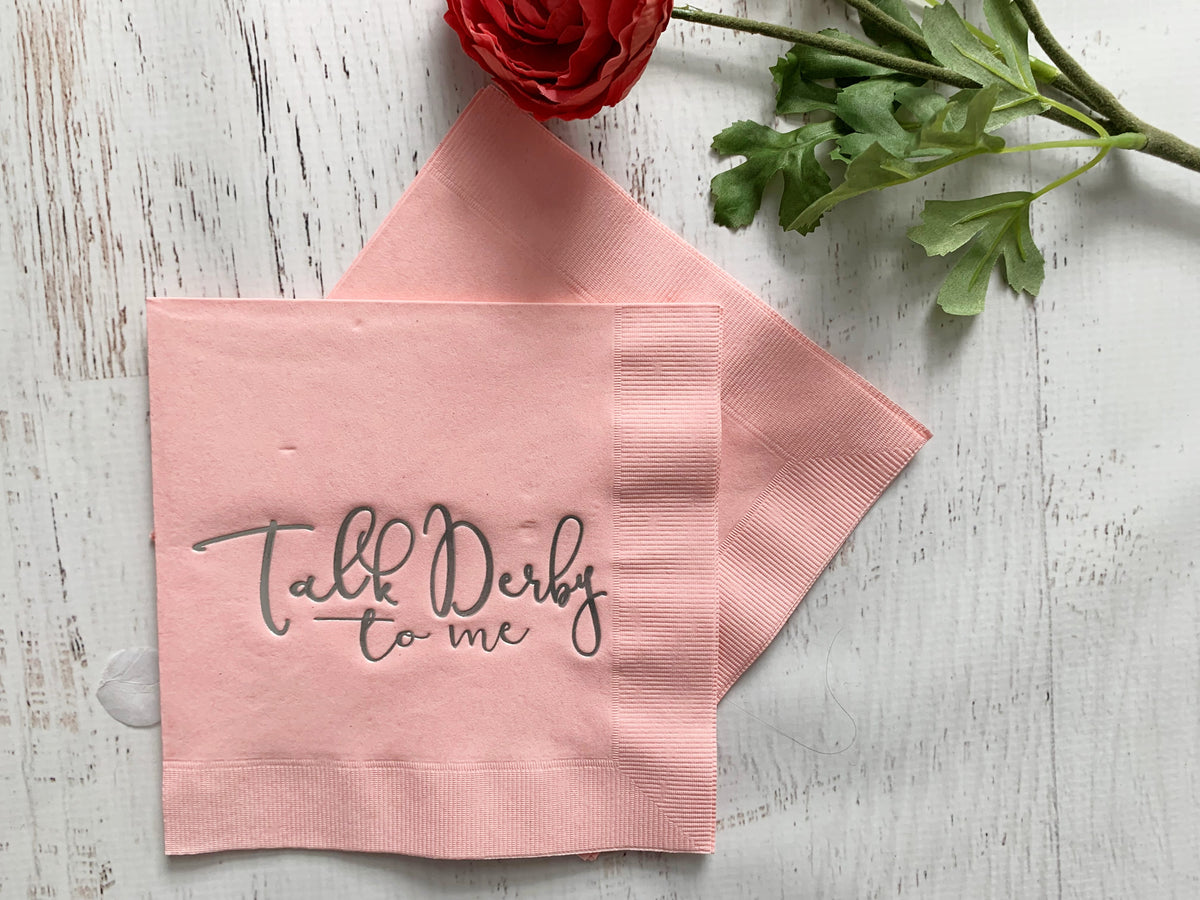 Pink napkin with 'Talk Derby to me' text on a wooden surface with flowers.