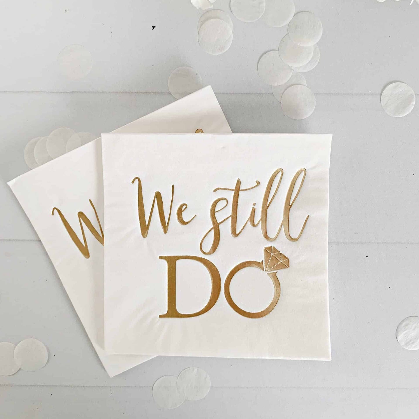 Cocktail napkin with 'We still do' text and a ring symbol on a white background with flowers.