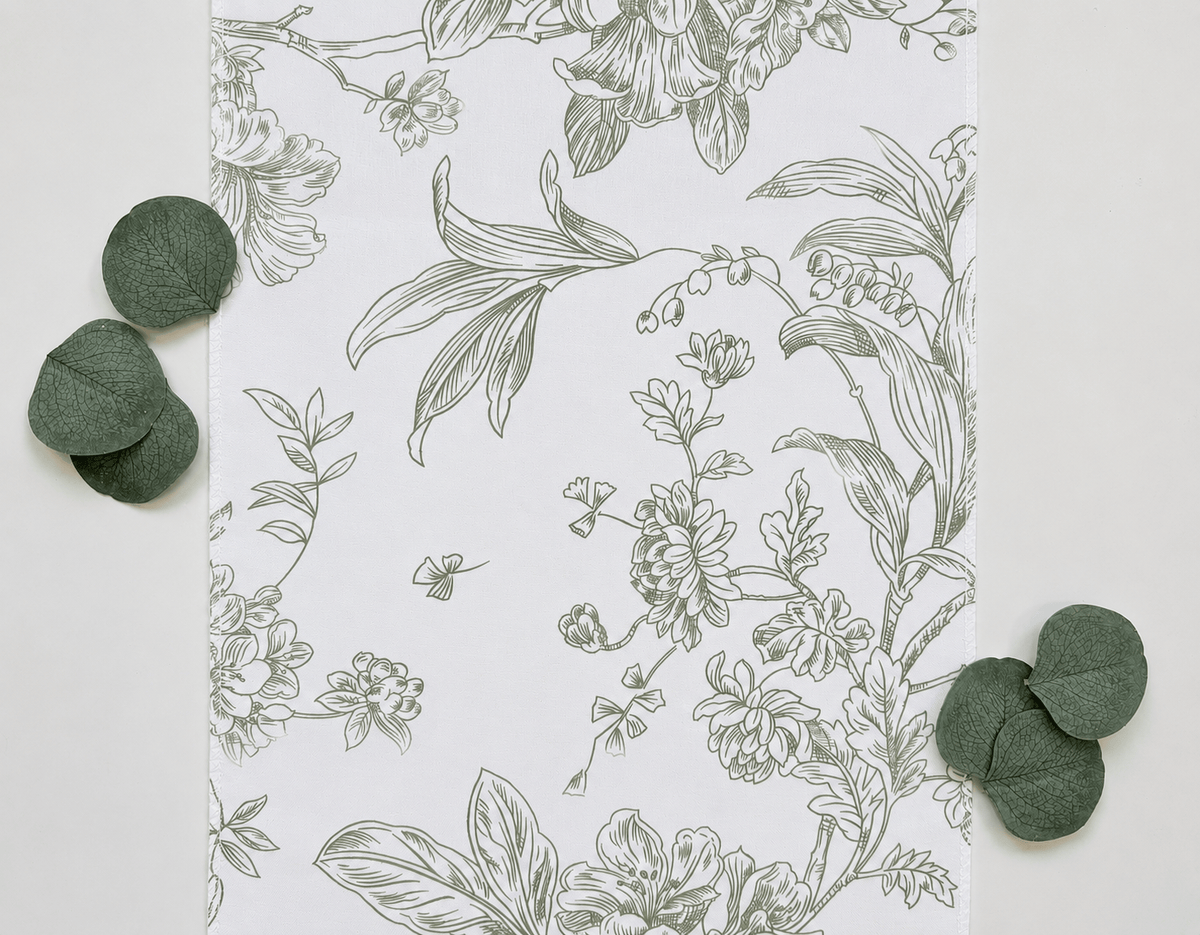 A flat-lay view of a white table runner featuring a delicate sage green botanical line drawing, styled with scattered green eucalyptus leaves on a light neutral background.