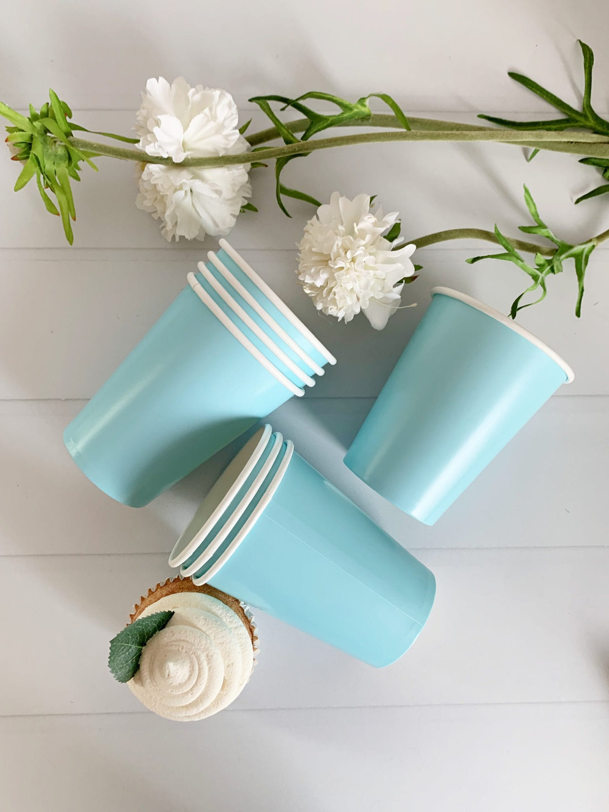 Two light blue paper cups on a light wooden surface with white flowers and green leaves.