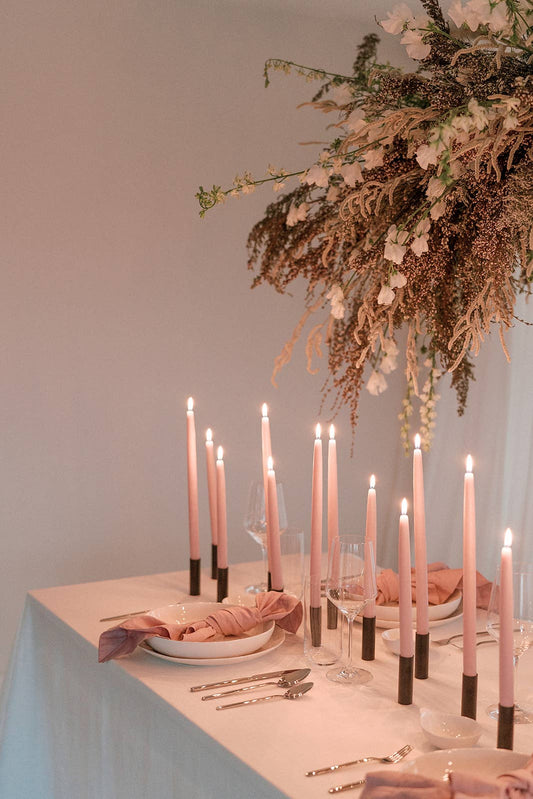 Progressive dinner table setting with candles, flowers, and cutlery on a neutral background - lots of slim pink taper candles gives the room a warm cozy glow.