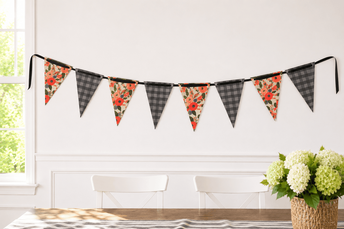 A string of triangular bunting flags hangs on a white wall in a bright room. The flags alternate between charcoal and grey plaid and floral prints with red, orange, and green tones reminiscent of Rifle Paper.