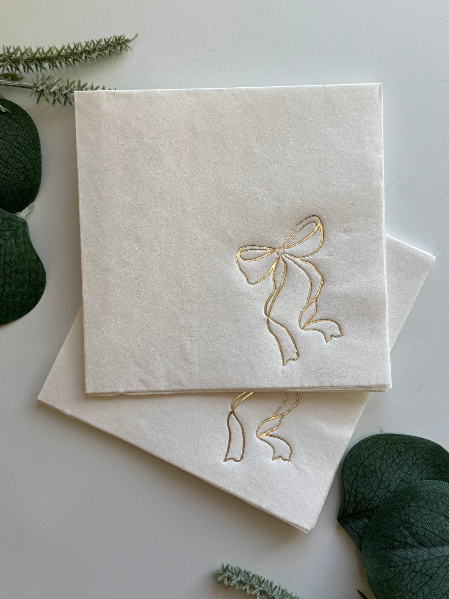 White napkin with gold bow design on a white background with greenery