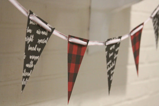A string of triangular bunting flags hanging across a white brick wall. The flags alternate between black with white script text and red-and-black buffalo plaid.