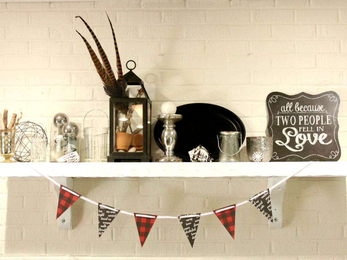 A styled holiday mantel display featuring a red and black buffalo plaid bunting banner hanging below a white shelf against a white brick wall. The shelf is decorated with a black lantern with feathers, silver and glass ornaments, a black plate, and a chalkboard sign that reads 'all because TWO PEOPLE LOVE IN Love'.