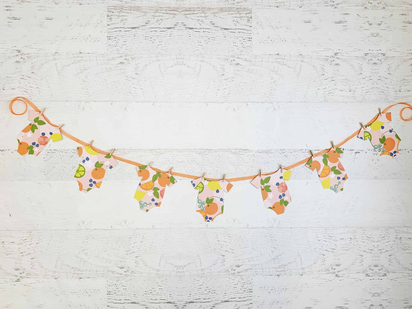A Little Cutie Citrus Onesie Banner featuring seven onesie-shaped cutouts in bright citrus fruit designs on a blush background, with an orange sheer organza cord and mini clothespins. Perfect for a Little Cutie-themed baby shower.