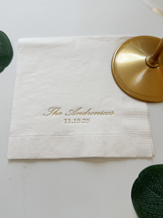 A white paper personalized wedding napkin with gold embossed text reading 'The Andromedas' and the date '11.15.