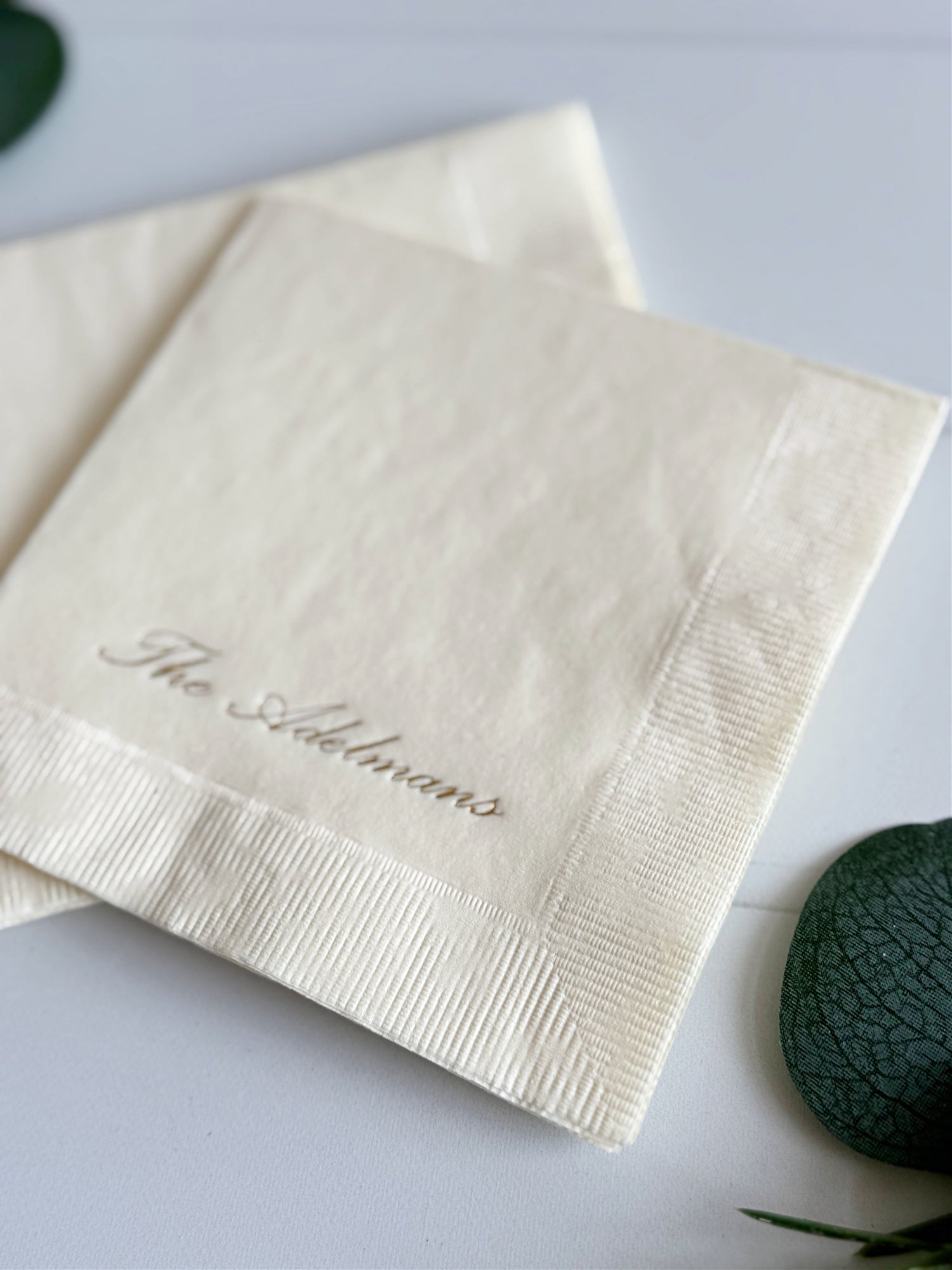 Two cream-colored paper napkins with a coined border, featuring the name 'The Adlmans' embossed in gold in a cursive font, displayed on a light surface with decorative green leaves.