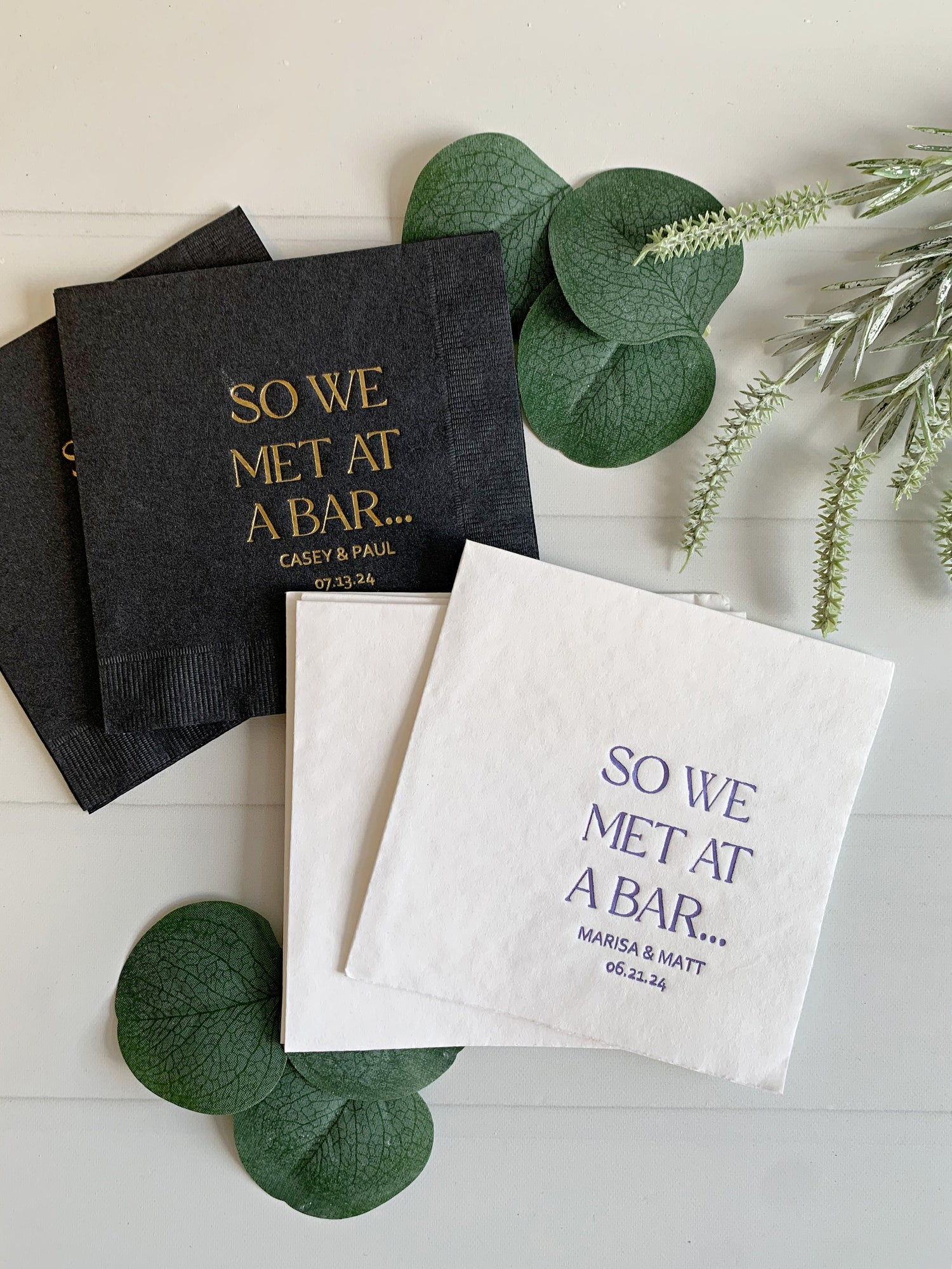 Two personalized paper napkins styled on a white wood background with green eucalyptus leaves. One napkin is dark charcoal gray with gold foil text reading 'SO WE MET AT A BAR.