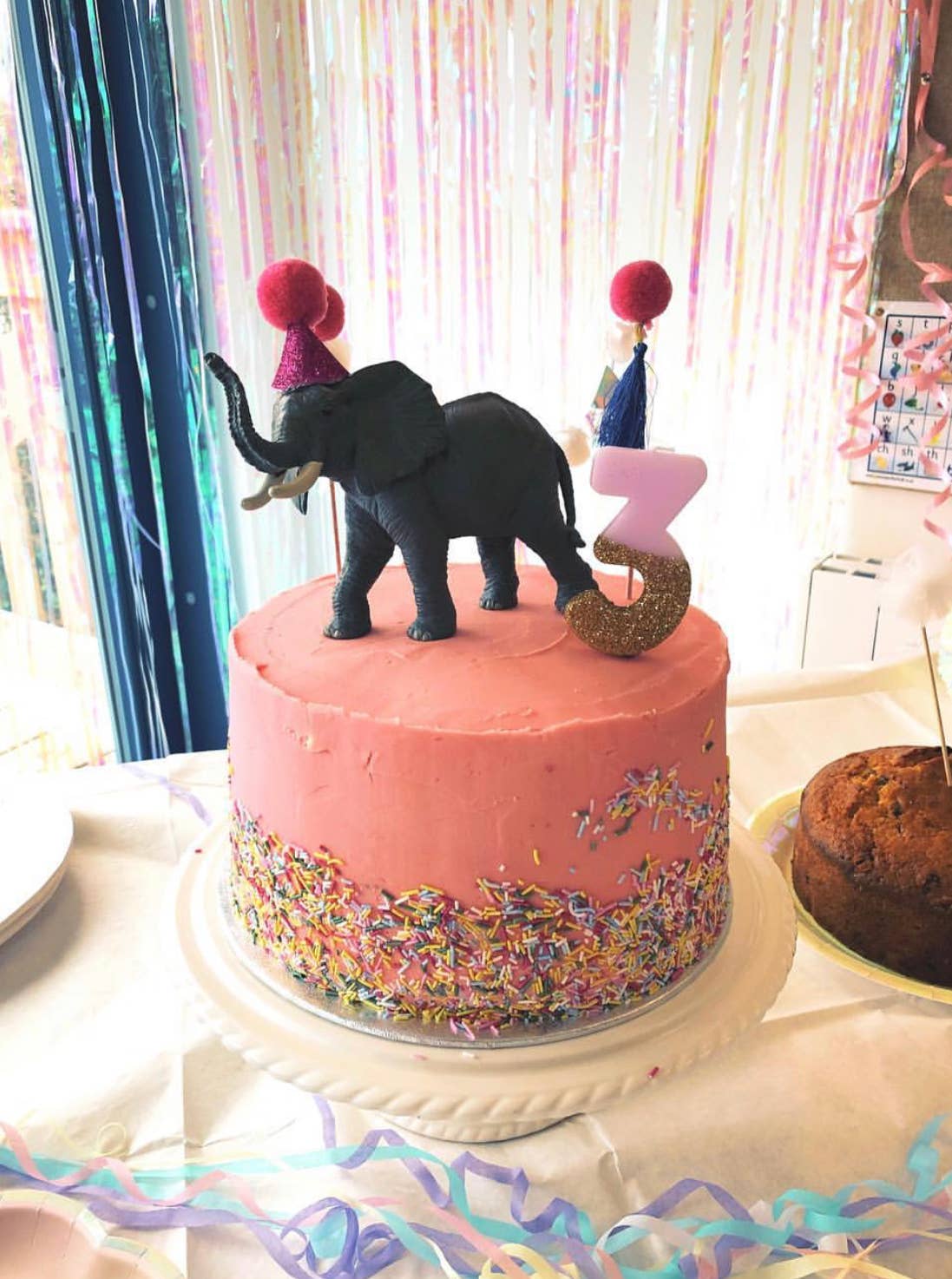 Pink birthday cake with elephant topper and number 3, surrounded by colorful decorations.