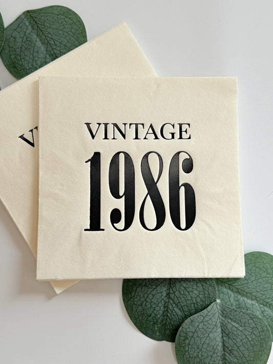 A square cream-colored cocktail napkin in 3 ply paper with the words 'VINTAGE 1986' printed in black serif lettering, styled with green leaves on a white surface.