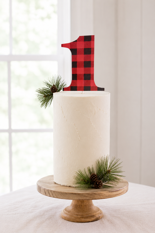 A white frosted cake on a round wooden pedestal stand, decorated with pine branches and a red-and-black buffalo plaid number 1 topper, set in front of a softly blurred window. A beautiful setup for a little lumberjack birthday party.