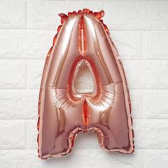 A metallic rose gold foil balloon shaped like the letter 'A', shown against a white brick wall background.