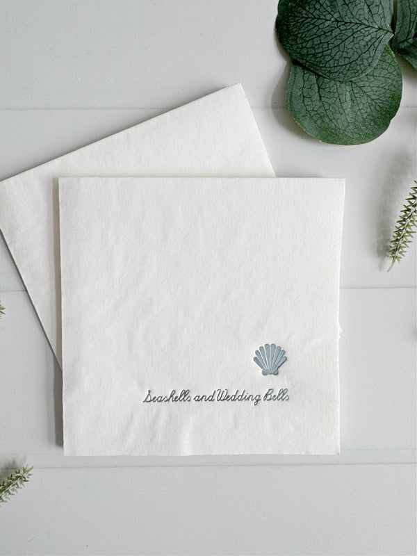 White napkin with shell design "seashells and wedding bells"  in ice blue foil text on a light gray background