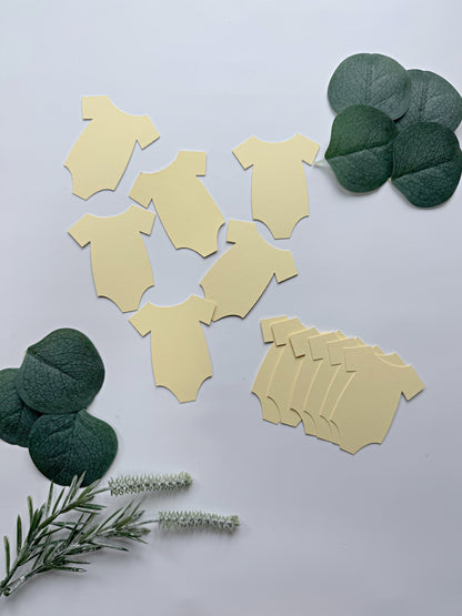 Pale yellow baby bodysuit-shaped party tags or confetti pieces arranged on a light gray background with decorative green eucalyptus leaves and rosemary sprigs.