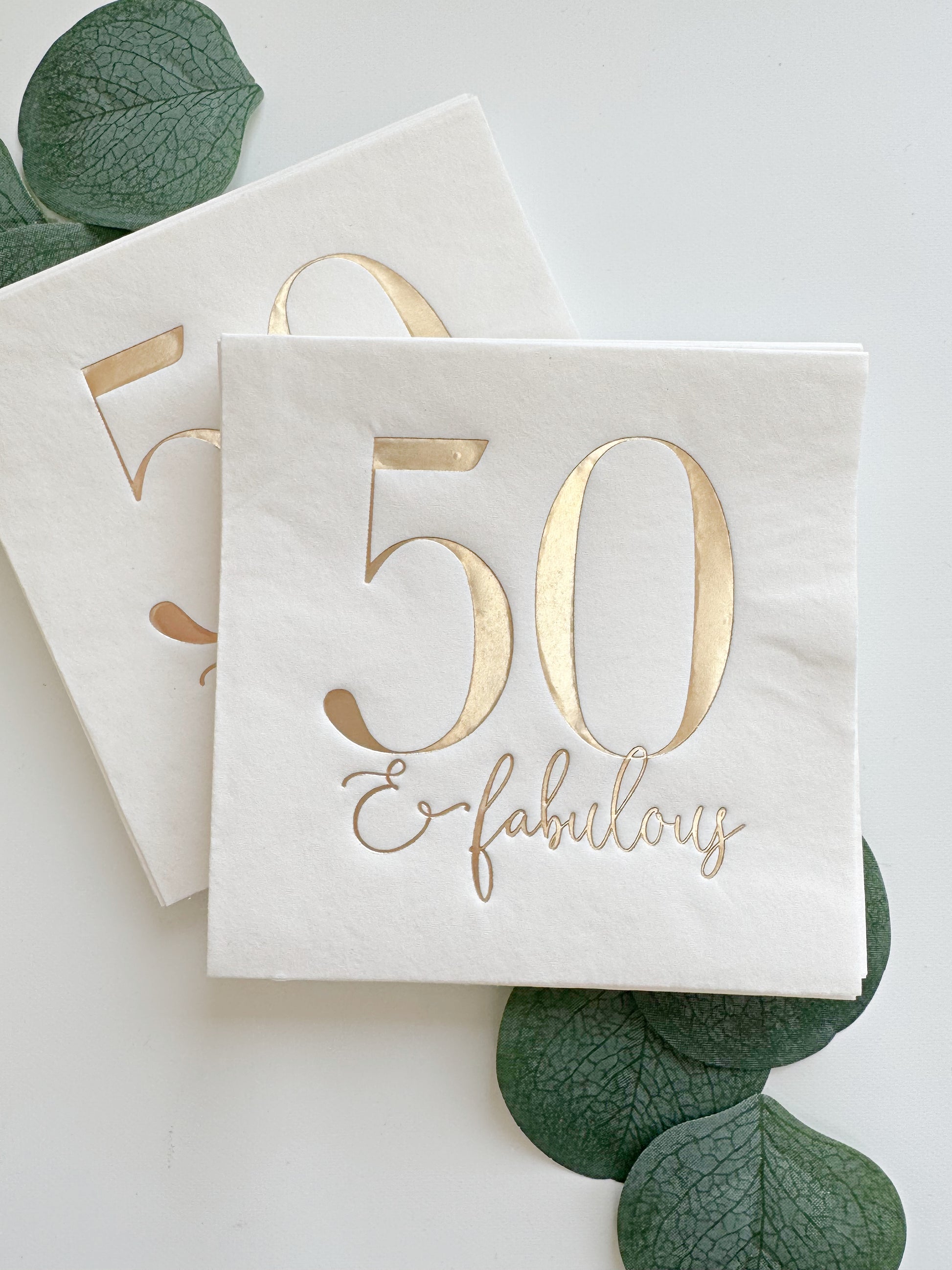 White napkins with gold foil '50 & fabulous' text on a white background with green leaves.