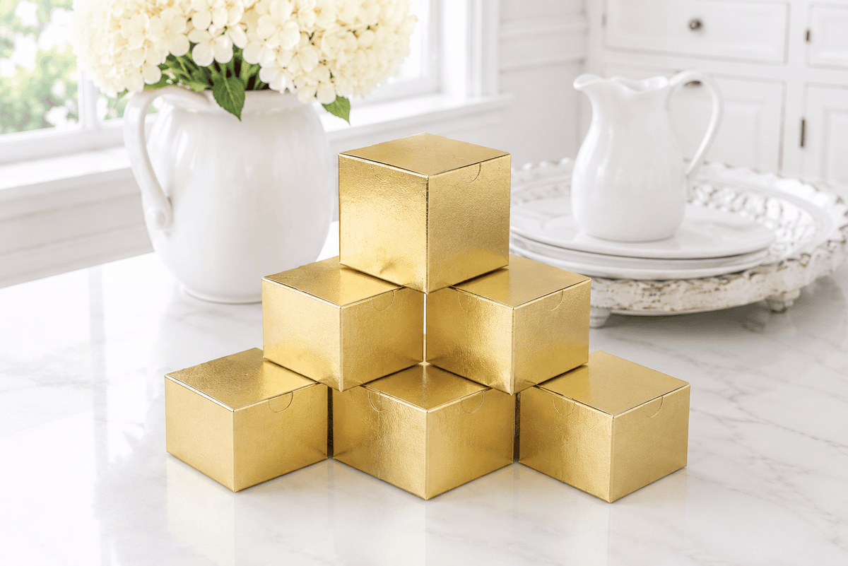 Seven metallic gold square favor boxes stacked in a pyramid arrangement on a white marble countertop, with a white ceramic pitcher and flowers in the background.
