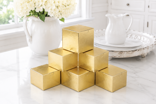 Seven metallic gold square favor boxes stacked in a pyramid arrangement on a white marble countertop, with a white ceramic pitcher and flowers in the background.
