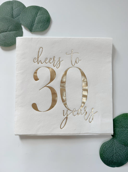 A square white paper napkin with metallic gold foil text reading 'Cheers to 30 years,' styled on a white surface with green leaf props in the corners.