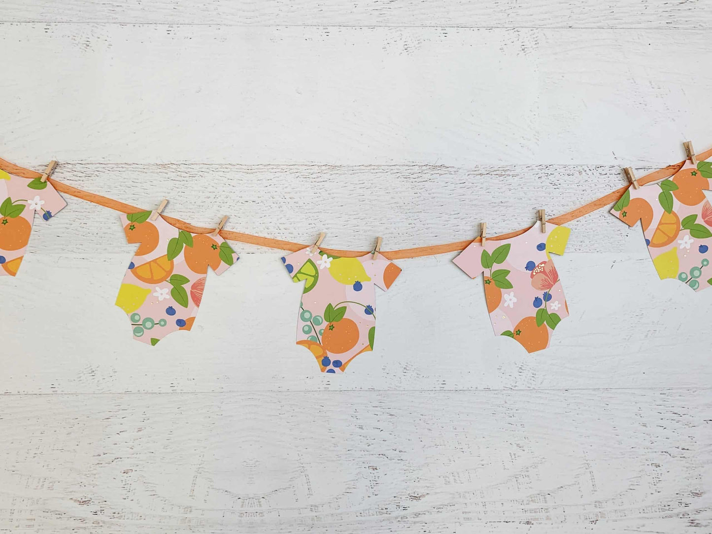 A Little Cutie Citrus Onesie Banner featuring seven onesie-shaped cutouts in bright citrus fruit designs on a blush background, with an orange sheer organza cord and mini clothespins. Perfect for a Little Cutie-themed baby shower.