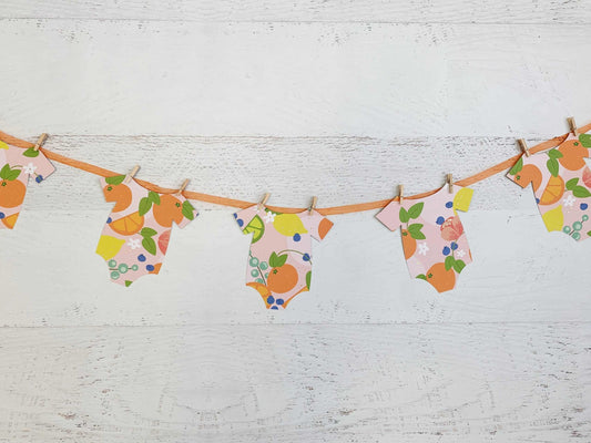 A Little Cutie Citrus Onesie Banner featuring seven onesie-shaped cutouts in bright citrus fruit designs on a blush background, with an orange sheer organza cord and mini clothespins. Perfect for a Little Cutie-themed baby shower.