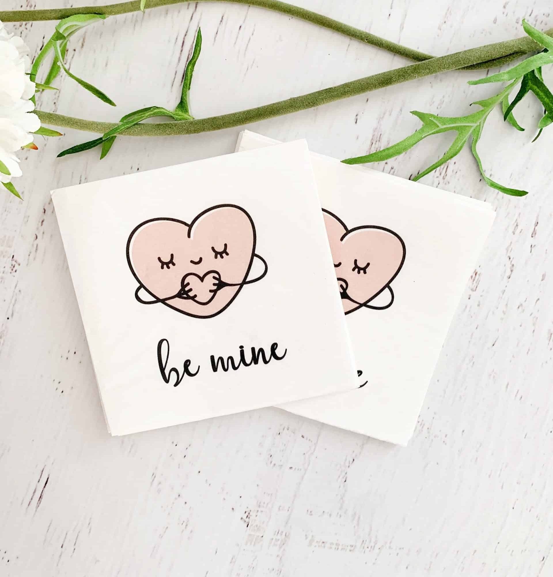 Sweet hugging hearts say "be mine" on these EnFete original cocktail napkins. White background with blush heart and black writing. Perfect for a Valentine treat, a Galentine's day cocktail, or under an afternoon snack.