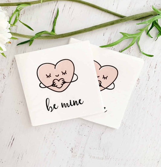 Sweet hugging hearts say "be mine" on these EnFete original cocktail napkins. White background with blush heart and black writing. Perfect for a Valentine treat, a Galentine's day cocktail, or under an afternoon snack.