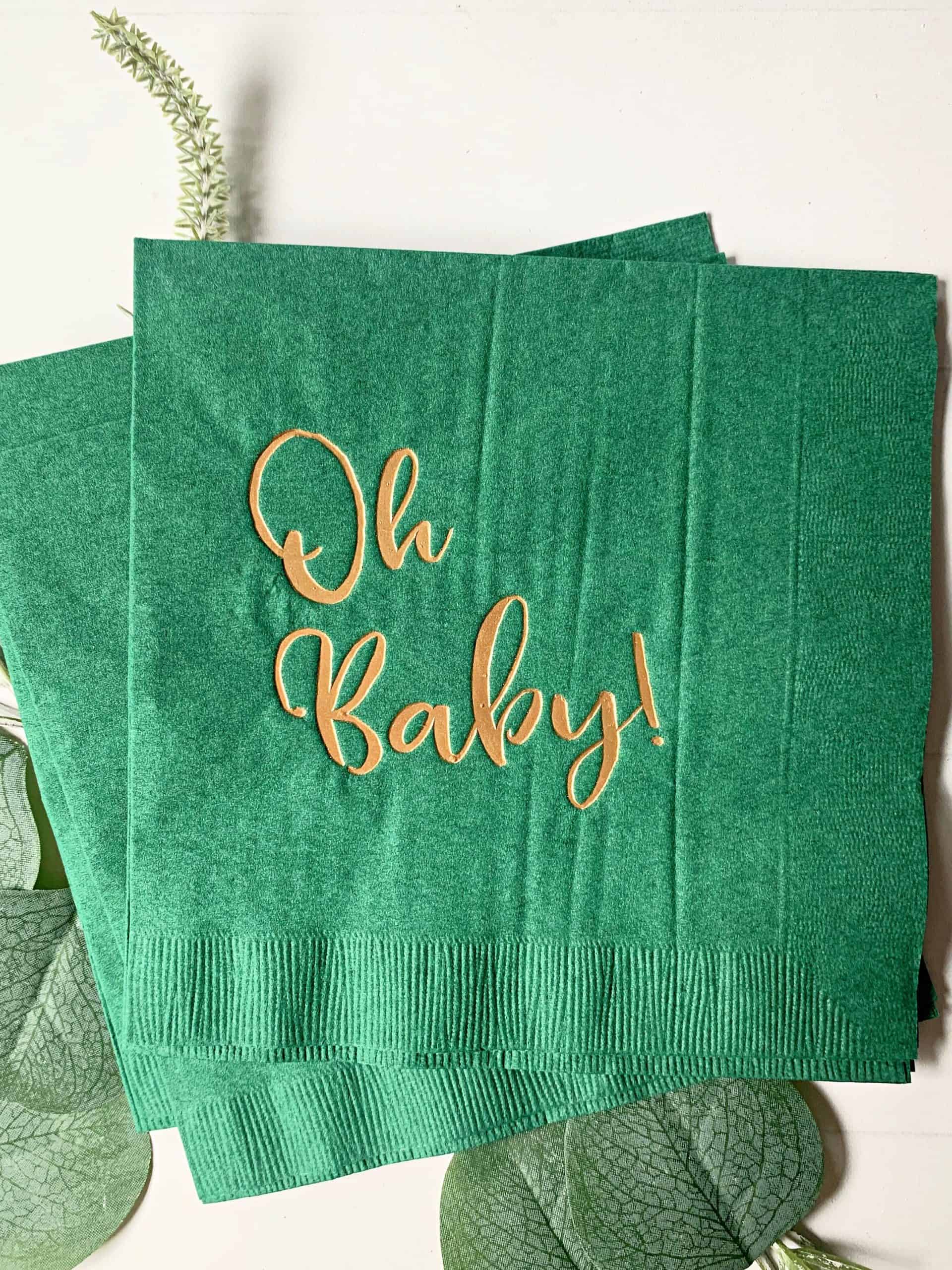 Picture of 16 'Oh Baby' luncheon napkins in gold foil script on forest green paper, perfect for a mountain adventure baby shower celebration.