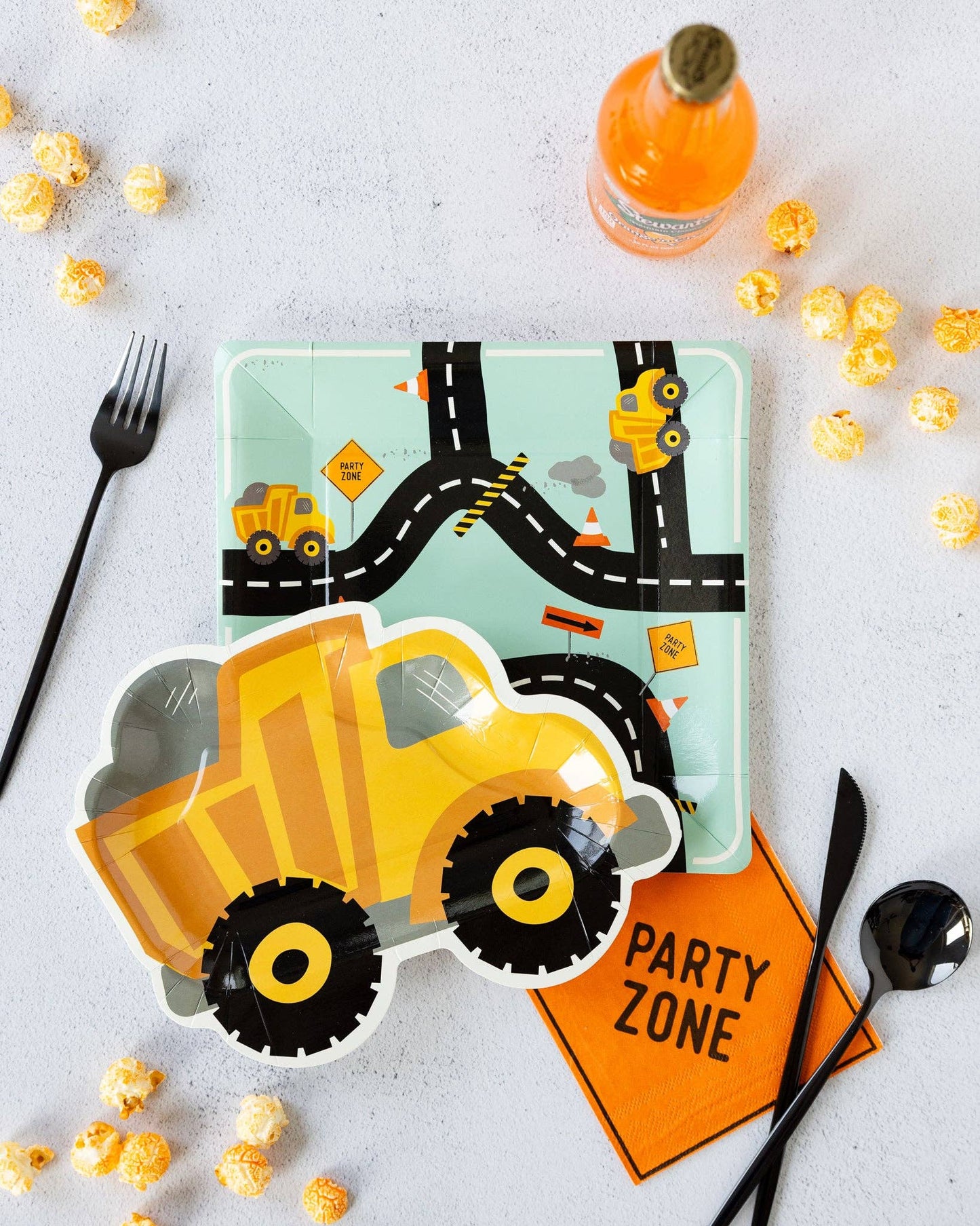 Children's plate with road design and dump truck, surrounded by popcorn and cutlery on a light surface.