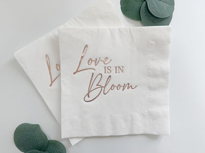 White paper luncheon napkin with 'Love is in Bloom' text in rose gold on a light gray background