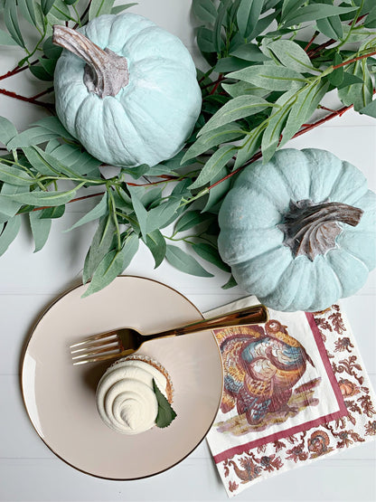 Two light blue pumpkins with green leaves on a white surface, next to a plate with a dessert and a decorative napkin.