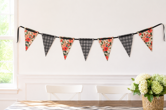 A string of triangular bunting flags hangs on a white wall above a dining table. The flags alternate between charcoal plaid and floral prints with coral red, orange, and green tones.