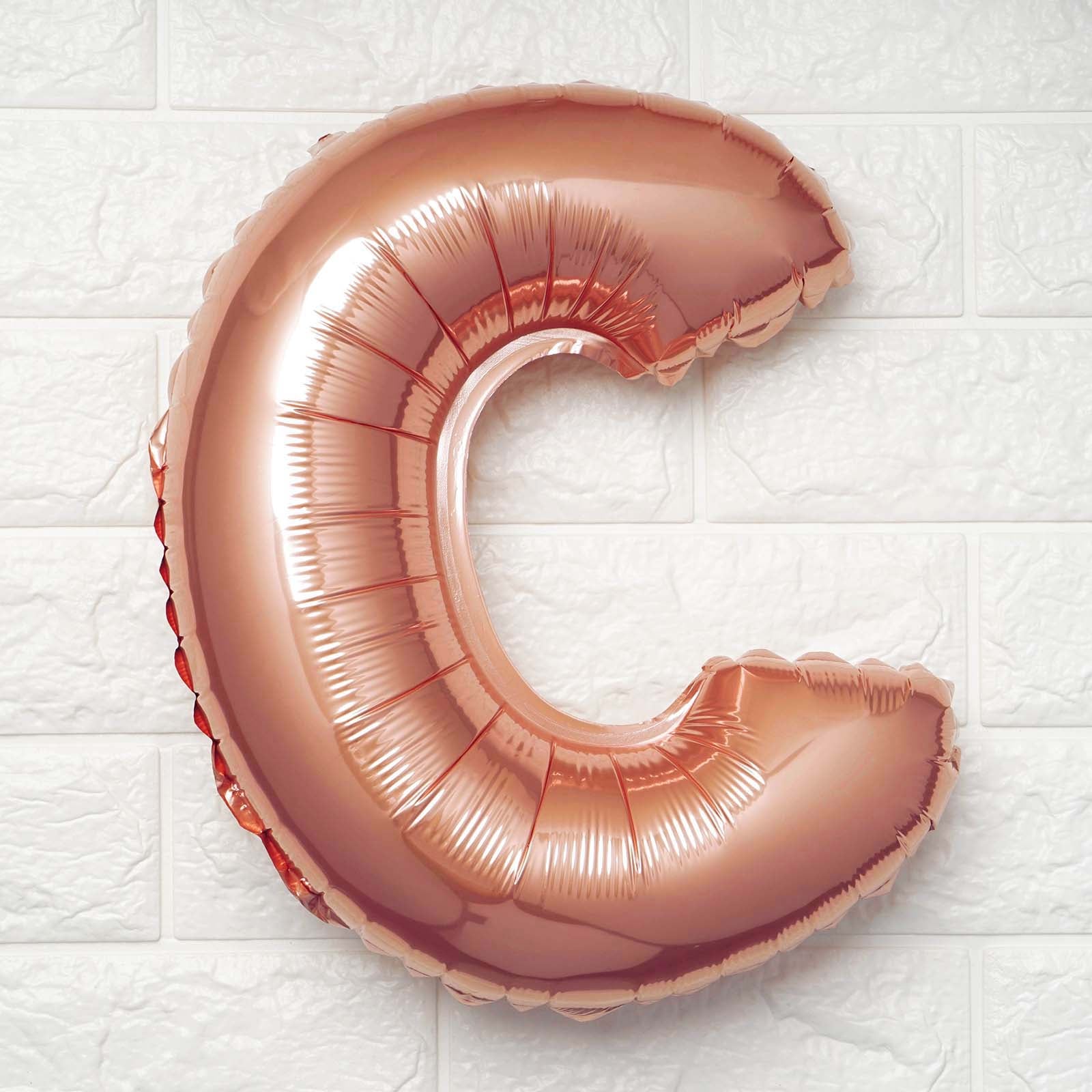 A metallic rose gold foil balloon shaped like the letter 'C', shown against a white textured brick wall background.
