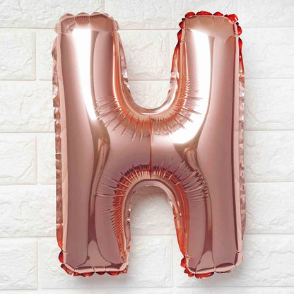 A metallic rose gold foil balloon shaped like the letter H, shown against a white textured brick wall background.