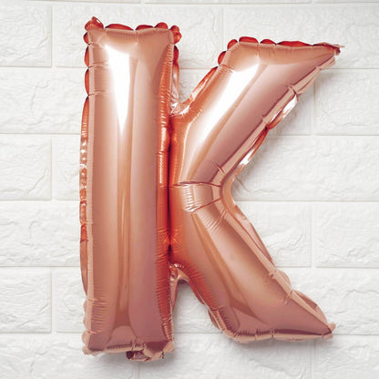 A metallic rose gold foil balloon shaped like the letter K, resting against a white textured brick wall background.
