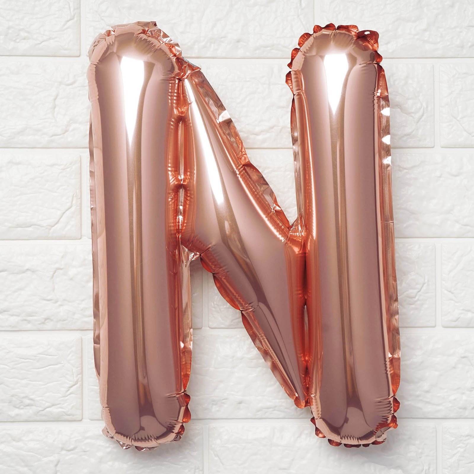 A metallic rose gold foil balloon shaped like the letter 'N', photographed against a white textured brick wall background.