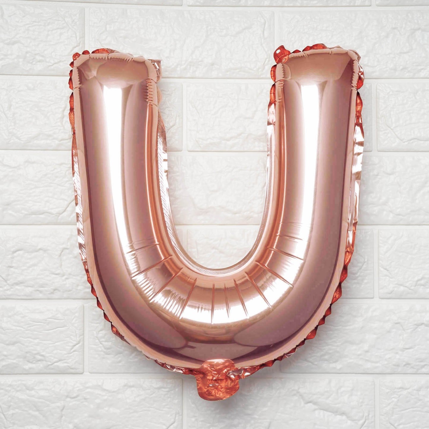 A metallic rose gold foil balloon shaped like the letter U, standing upright against a white textured brick wall.