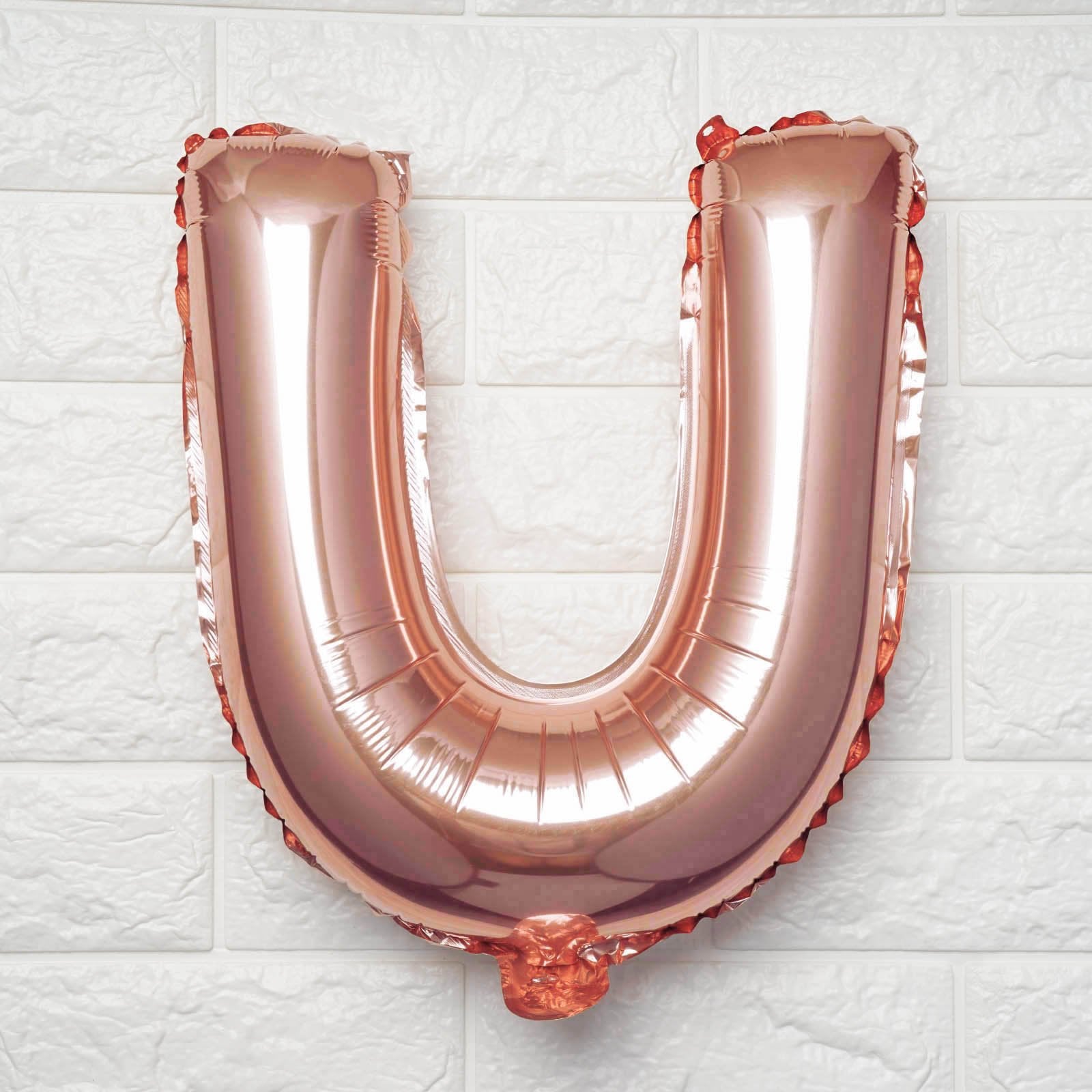 A metallic rose gold foil balloon shaped like the letter U, standing upright against a white textured brick wall.