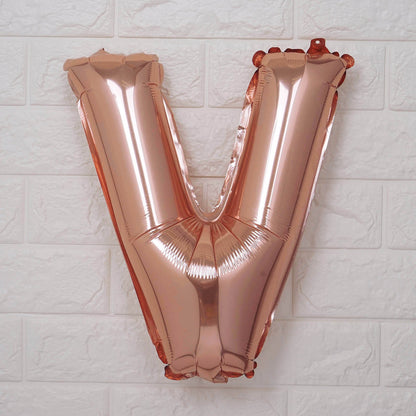 A metallic rose gold foil balloon shaped like the letter 'V', mounted against a white textured brick wall.