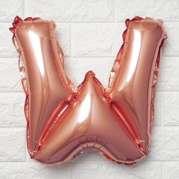 A metallic rose gold foil balloon shaped like the letter 'W', shown against a white textured brick wall background.