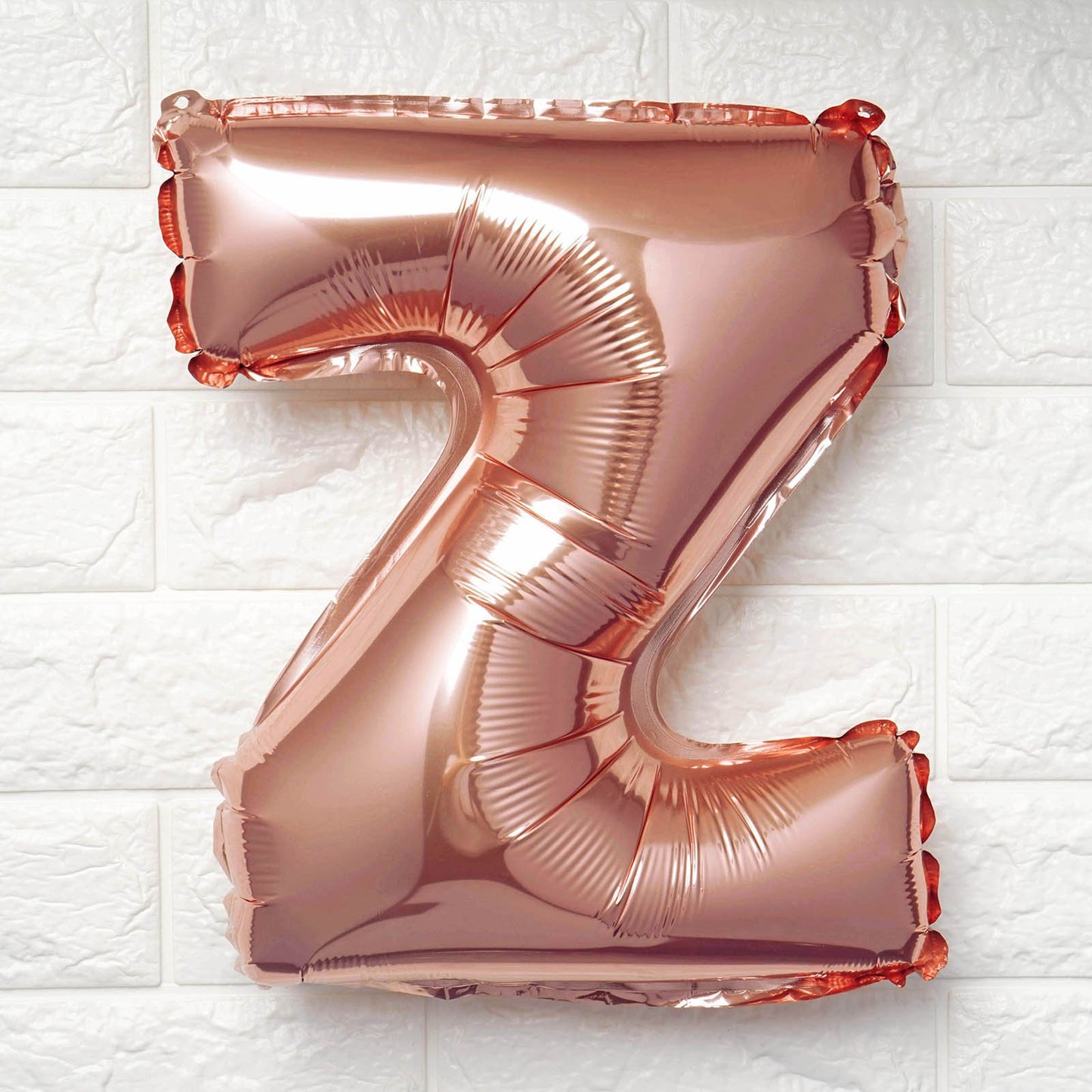 A metallic rose gold foil balloon shaped like the letter 'Z', floating against a white textured brick wall background.